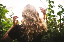 Blondynka z falowanymi średnioporowatymi włosami stoi w zielonym ogrodzie.