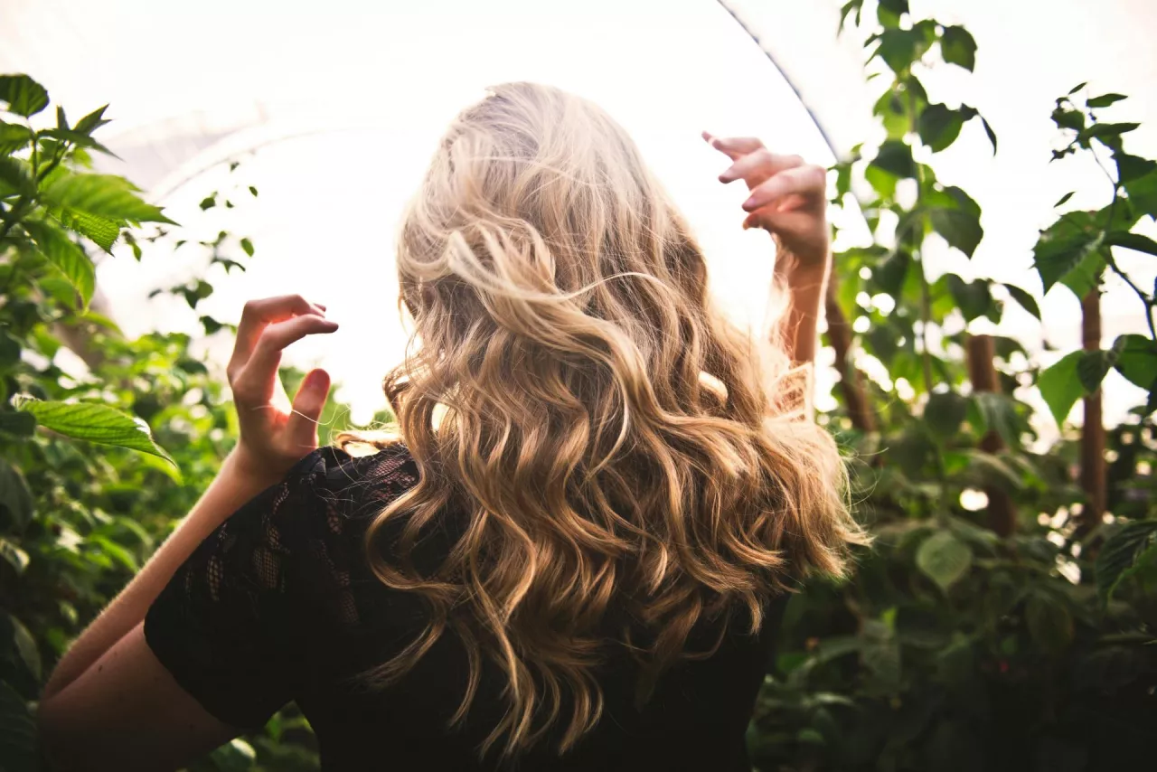 Blondynka z falowanymi średnioporowatymi włosami stoi w zielonym ogrodzie.